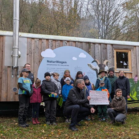 Eine Gruppe Menschen steht von einem Bauwagen. Im Vordergrund wird ein Fördercheck gezeigt.