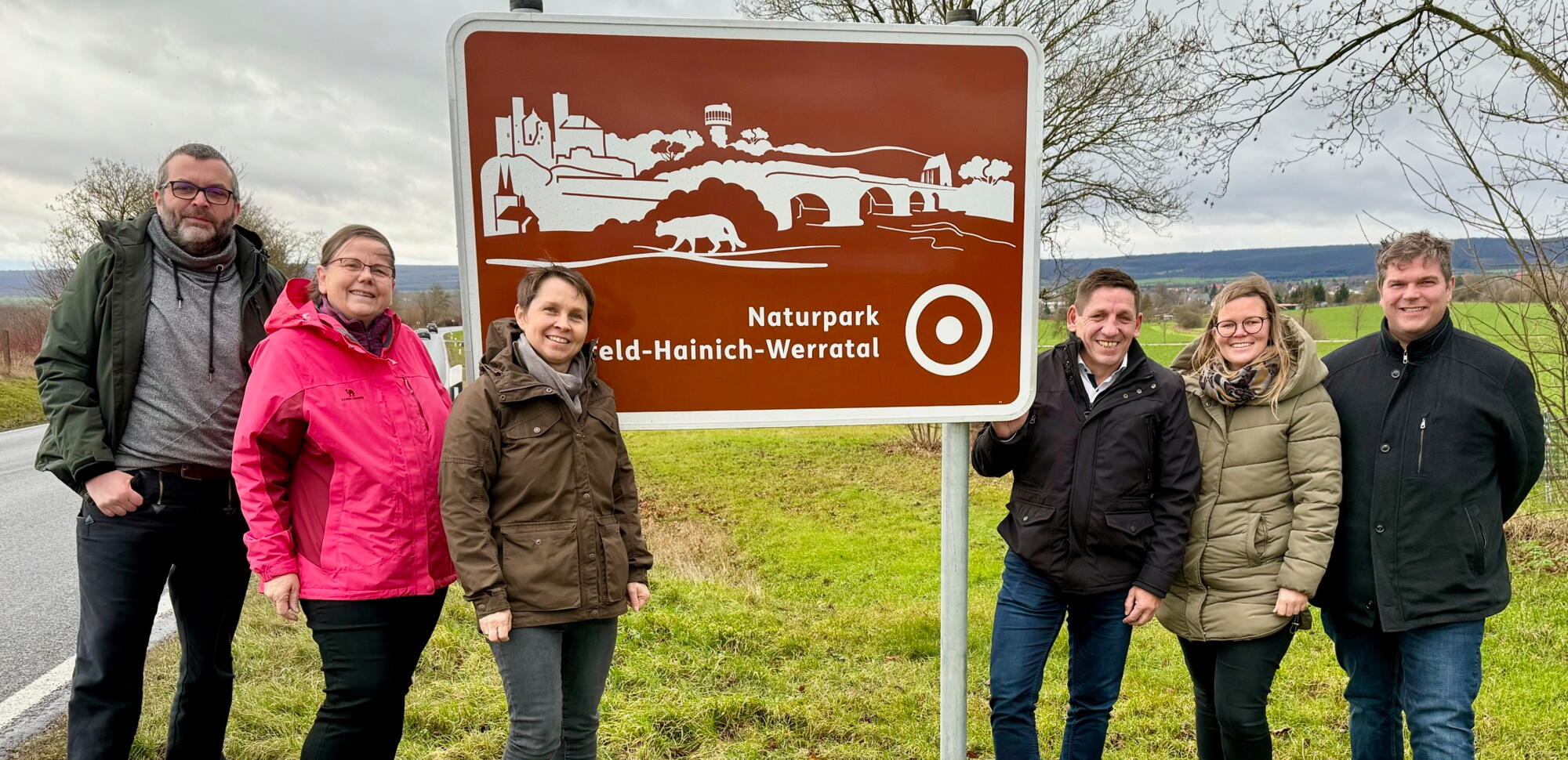 Gruppe von sechs Personen steht neben einer neuen braunen Willkommenstafel des Naturparks Eichsfeld-Hainich-Werratal. Die Tafel zeigt stilisierte Symbole des Naturparks, darunter Gebäude, Brücken und einen Bären. Im Hintergrund ist eine offene Wiesenlands