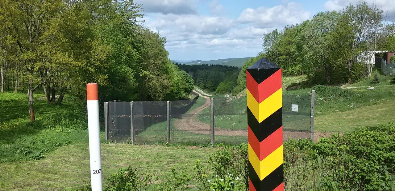 Im Vordergrund ist eine schwarz-rot-gelbe Grenzsäule zu sehen, dahinter der ehemalige Grenzzaun an der innerdeutschen Grenze.
