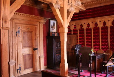 Historisch, mittelalterlicher Raum. Große Holztür mit historischen Elementen. Wände im Mix aus Holz und roter Tapete. Holzsäulen im Raum mit Schnitzereien. Blick auf eine Sitzgruppe mit Tisch und Stühlen im historischen Stil.