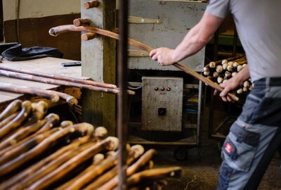 Werkstattszene bei der Herstellung von Wanderst?cken aus Holz. Eine Person bearbeitet lange Holzst?be an einer Maschine. Im Vordergrund liegen zahlreiche gebogene Wanderst?cke, w?hrend im Hintergrund weitere Materialien und Werkzeuge zu sehen sind.