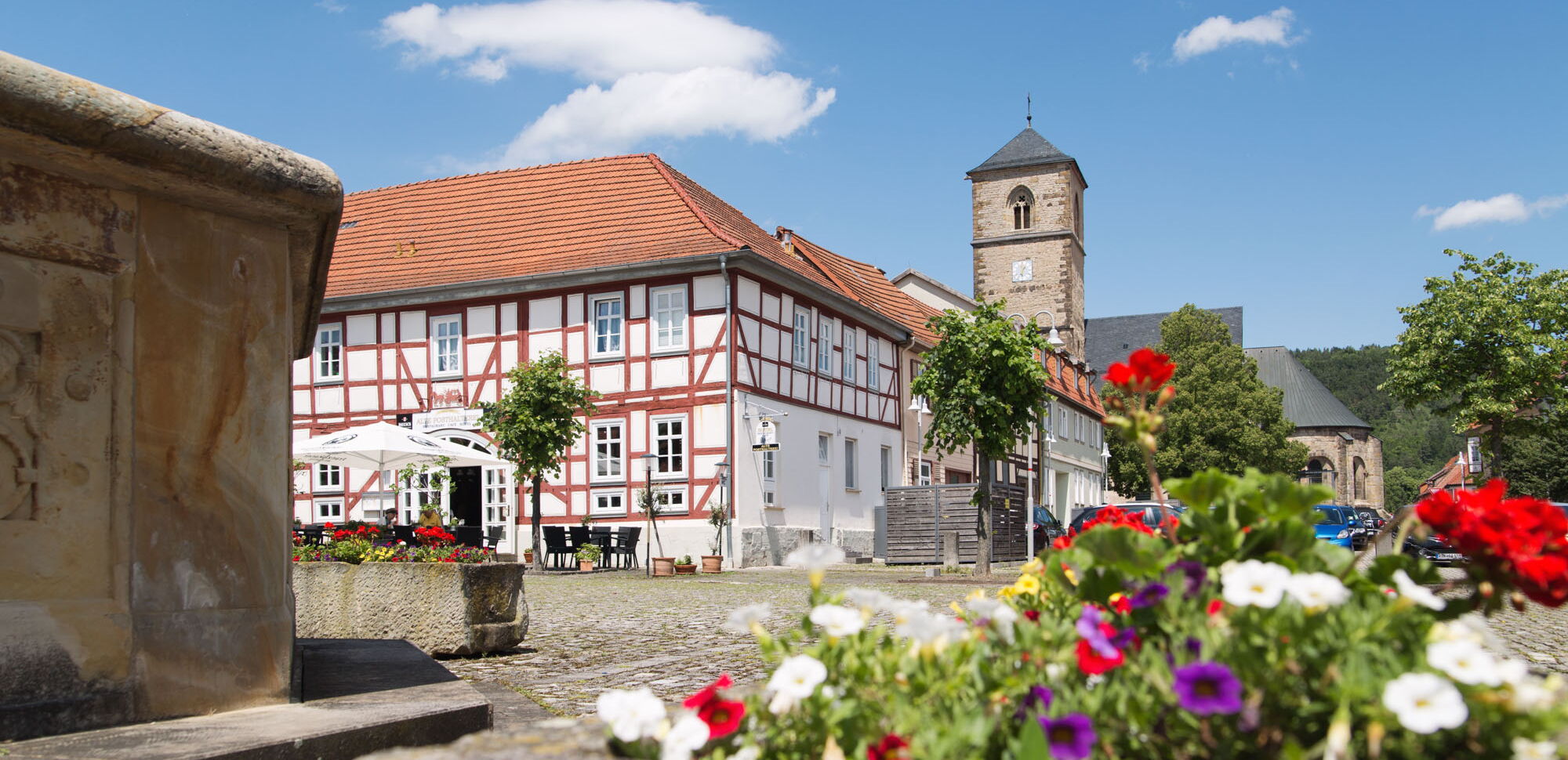 Markt in Creuzburg mit Brunnen und Blumendetail im Vordergrund. Im Hintergrund sind Kirche und Fachwerkhäuser zu sehen.