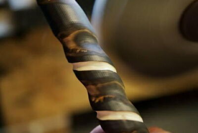 Nahaufnahme eines handgefertigten Wanderstocks aus dunklem Holz mit hellen Ringverzierungen. Im Hintergrund ist unscharf eine Schleifmaschine in einer Werkstatt zu sehen. Eine Hand h?lt den Stock w?hrend der Bearbeitung fest.