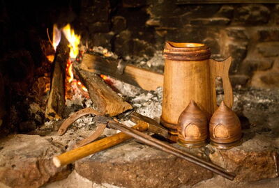 Blick in einen Steinernen Kamin. Feuer brennt, drei Holzspalten sind eingelegt. Davor Schmiedewerkzeug mit einem Hufeisen, einem Holzkrug und zwei Keramikbechern.